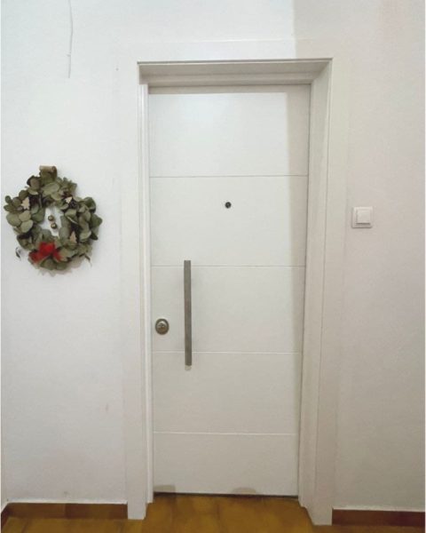 Puerta acorazada blanca con fresados horizontales y tirador vertical en un recibidor