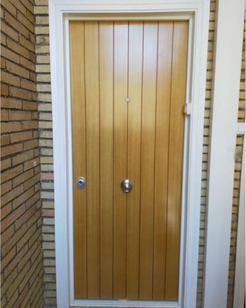 Puerta acorazada en madera con lamas verticales y dos bombines en una entrada de ladrillo