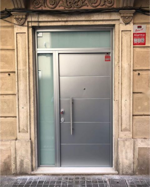 Puerta acorazada gris con fijo lateral y montante acristalados en un portal de piedra