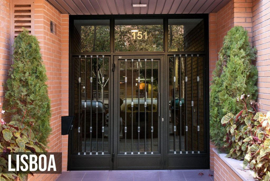 Puerta de portal modelo Lisboa, con hoja central y dos fijos laterales de barrotes rectos con remaches sobre vidrio, y montante de vidrio encima