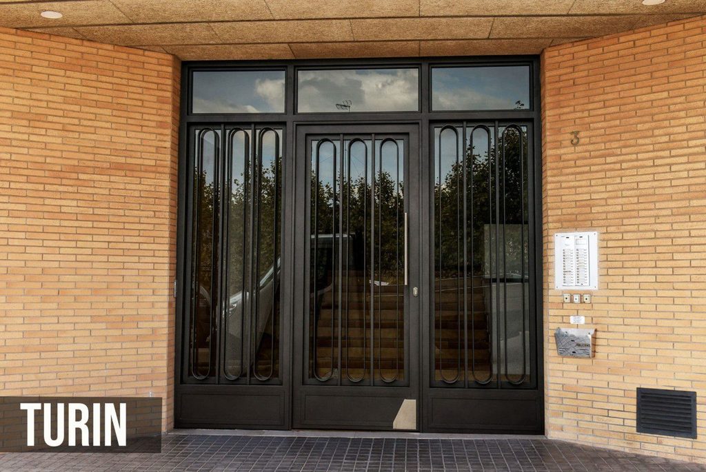 Puerta de portal modelo Turín, con hoja central y dos fijos laterales de barrotes verticales rematados en arcos de medio punto, y montante de vidrio encima